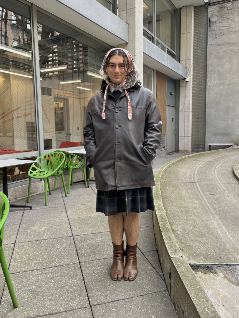 Art school student Sean Marner stands in a courtyard wearing a bonnet, leather jacket, plaid skirt, and brown leather tabi boots.
