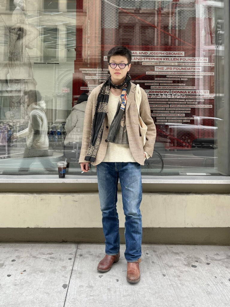 Art school student Zhuolun Lin takes a smoke break while wearing brown boots, jeans, a beige jacket, and a patterned scarf.