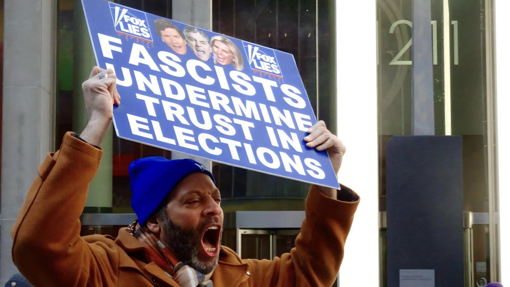 A man wearing a blue hat, flannel scarf, and brown jacket screams out in protest., He holds a blue sign with the faces of Tucker Carlson, Sean Hannity, and Laura Ingraham that reads in white letters, “FASCISTS UNDERMINE TRUST IN ELECTIONS” LYING NORMAL.”
