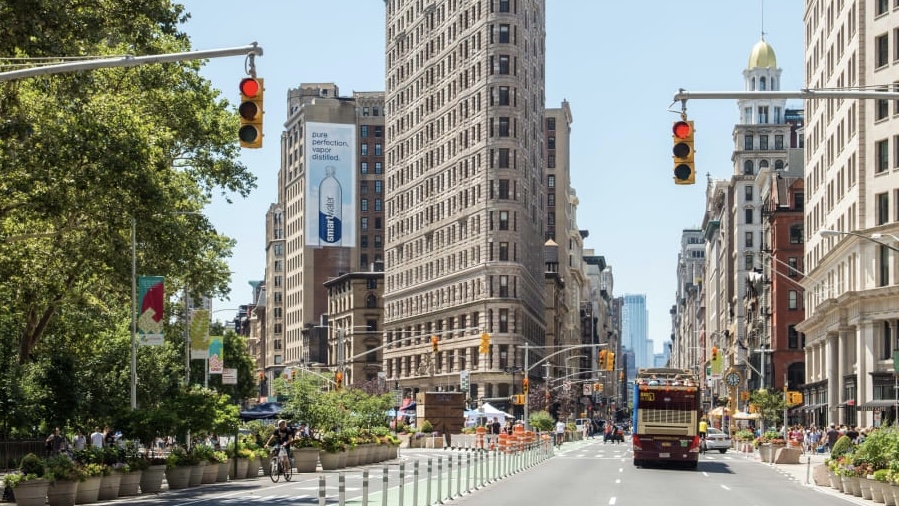 The flatiron building, a large, triangular building standing at the center of an intersection, sits next to a park.
