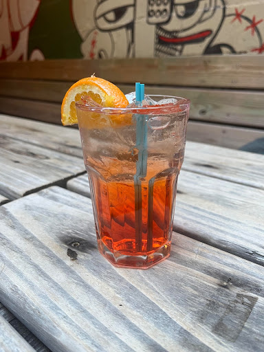 One aperol spritz outside on a wooden table from Sweet and Vicious. It has an orange ombre color and is garnished with an orange slice. 
