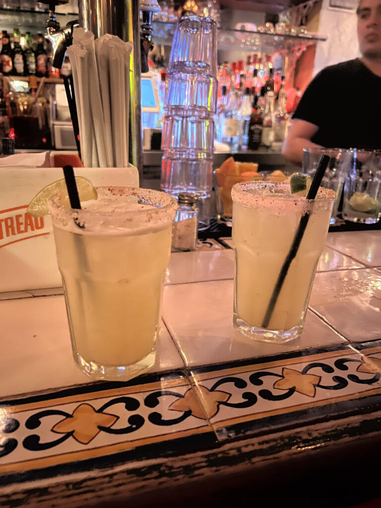 Two margaritas on the rocks with salt around the rim from Yuca bar. They are placed on a tile bar that has a blue and yellow pattern around the edge. 