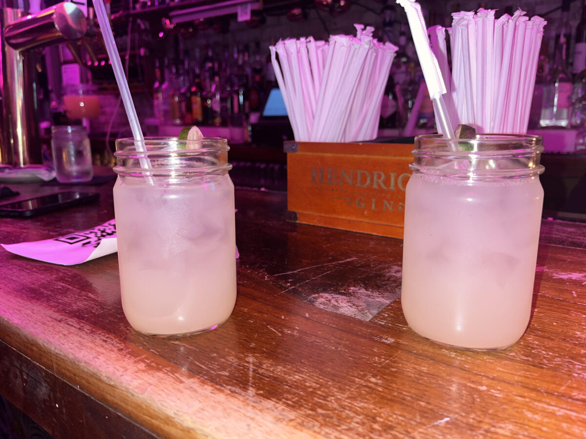 Two margaritas in mason jars with plastic straws from POCO. They are lit by purple LED lights and positioned on a wooden bar. 