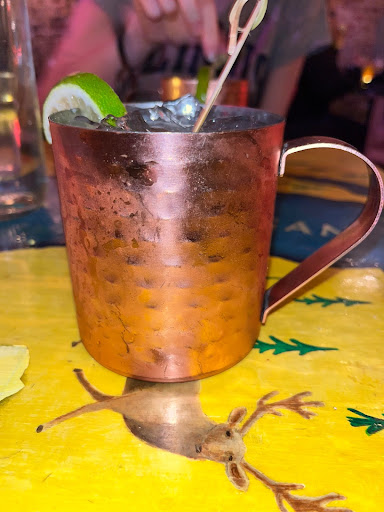 One Mexican mule in a tin cup from Grey Dog, with a lime and cherry garnish. They are placed on a yellow table that has drawings of deers and trees. 