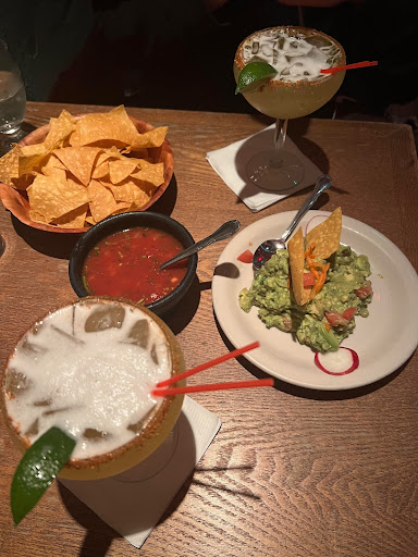 Two spicy margaritas paired with chips, guacamole, and salsa from El Cantinero. They are dimly lit and placed on a wooden table. 