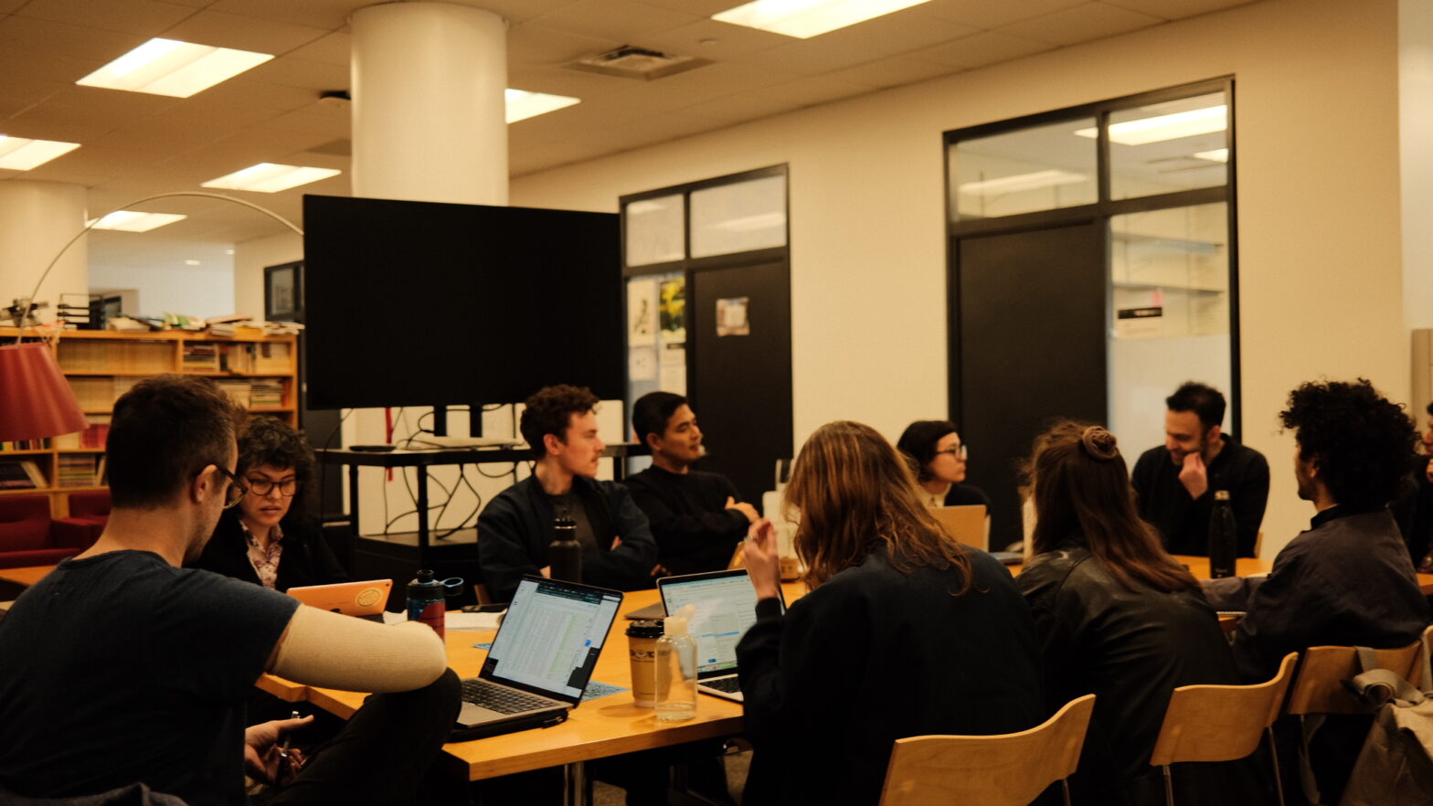 an image of nine people gathered around a table in a lounge speaking to each other