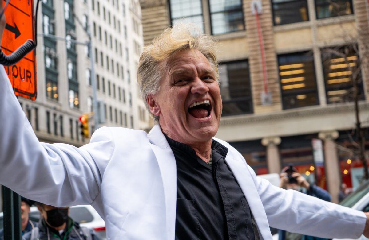 A man gives a speech in a white suit and black dress shirt during The New Schools part time faculty strike. He stands on Fifth Avenue in front of the University Center, the Welcome Center and Parsons building visible behind him