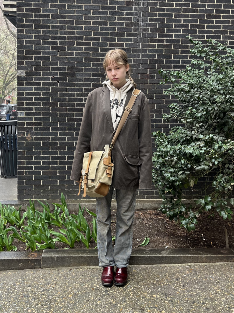 First-year journalism student wears a brown coat, beige hoodie, and pinstripe pants with a messenger bag and red clogs while standing in front of a school building.