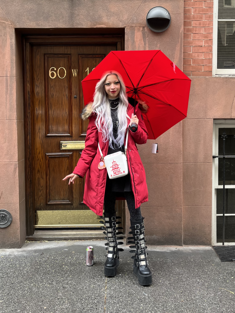 Second-year philosophy student wears a red coat, tall black boots, and has silver hair while holding a red umbrella and standing in front of a brownstone on Fifth Ave.