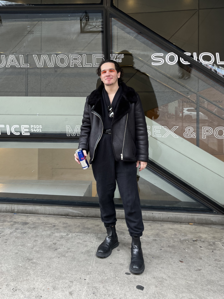 Third-year fashion design student wears black boots, a black jumpsuit, and a black leather jacket with bright orange eye makeup while sipping a redbull outside of the University Center on Fifth Ave.