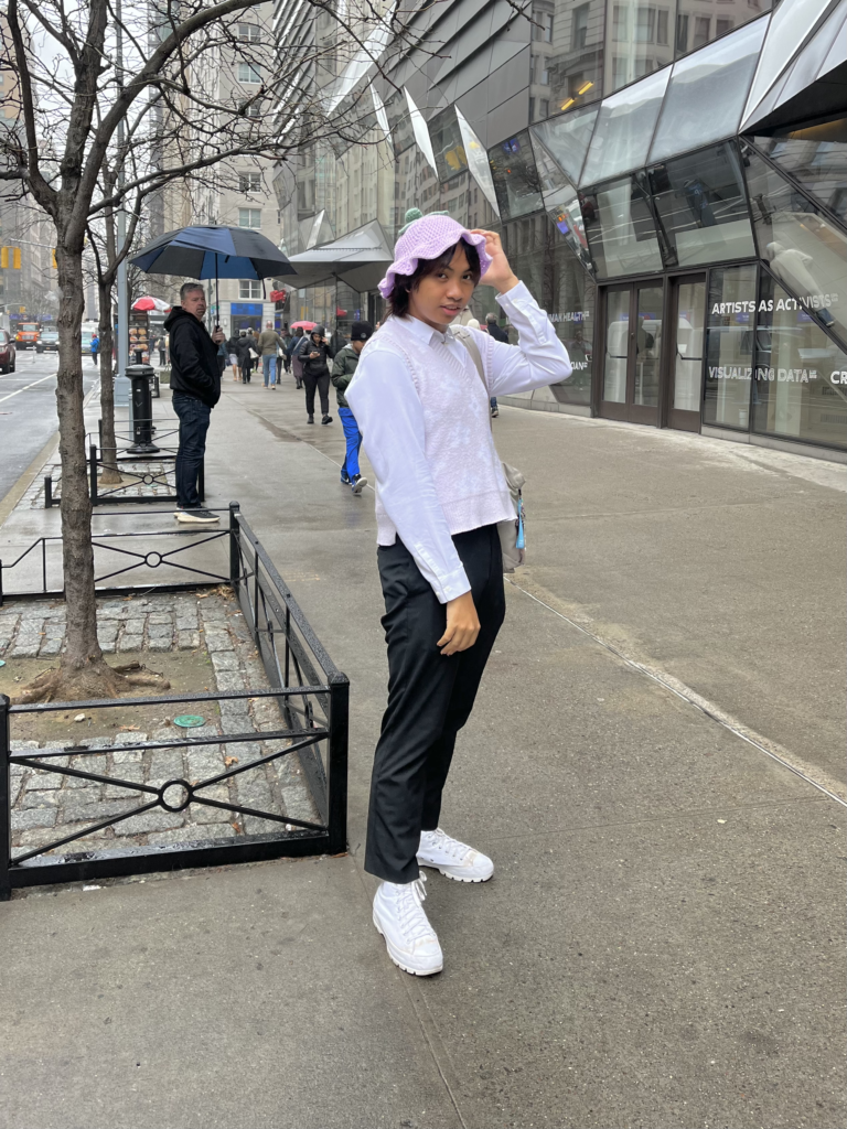 Fourth-year fashion design student wears white sneakers, black trousers, a white button-down over a pastel sweater vest, and a purple crochet hat while posing on Fifth Ave.
