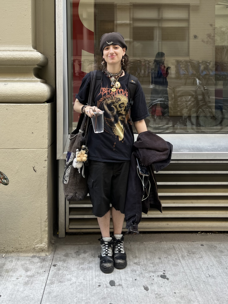 Second-year fine arts student wears black boots, black shorts, and a black graphic t-shirt with layered beaded necklaces while standing on Fifth Avenue.