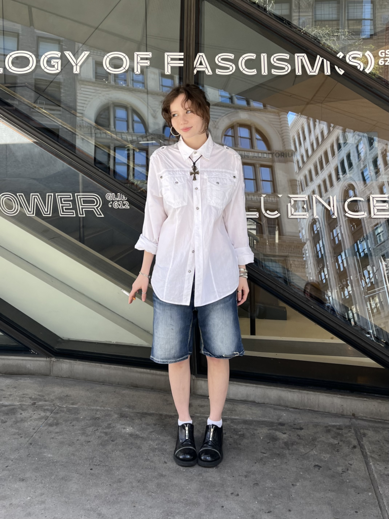 First-year fashion design wears black boots, jean shorts, a white button-up, and a silver cross necklace while on her smoke break.