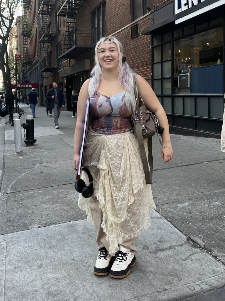 Student wears platform sneakers, a white lace skirt, graphic corset, and has purple hair while standing on a street corner.