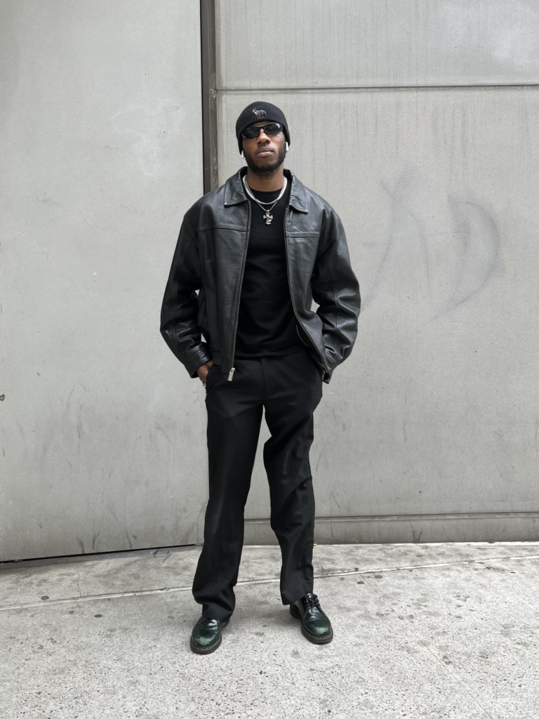 Student wears all-black boots, slacks, leather jacket, beanie, and sunglasses with silver jewelry while standing in front of a gray building.