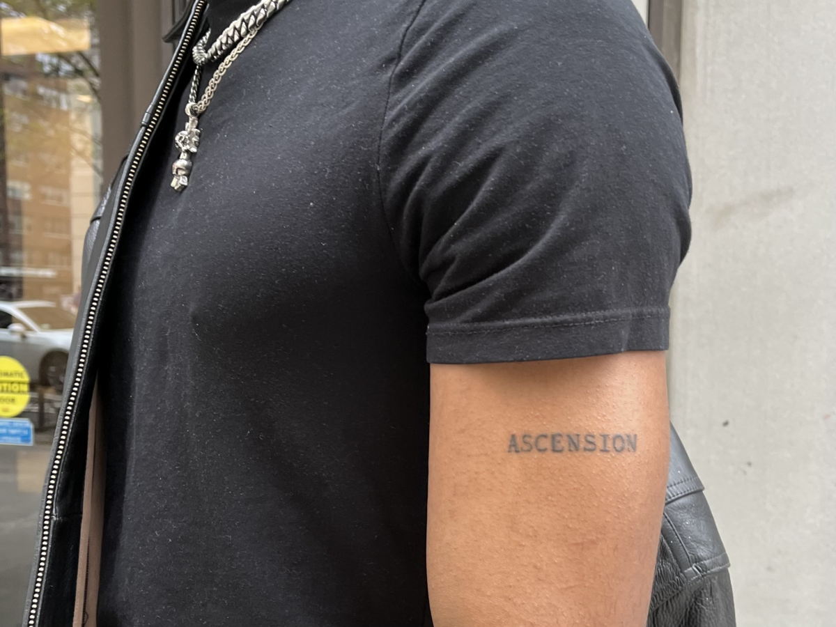 Torso dressed in a black short sleeved t-shirt with the word “ASCENSION” tattooed in all caps on bicep.