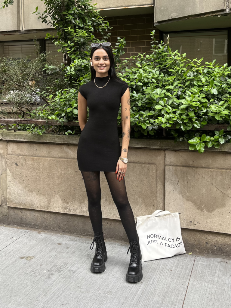 Student wears a short sleeved black dress, sunglasses on top of her head, black tights, boots, gold jewelry, and her left arm is covered in patchwork-style tattoos. She stands in front of green bushes and her tote bag that sits on the ground says “normalcy is just a facade.”