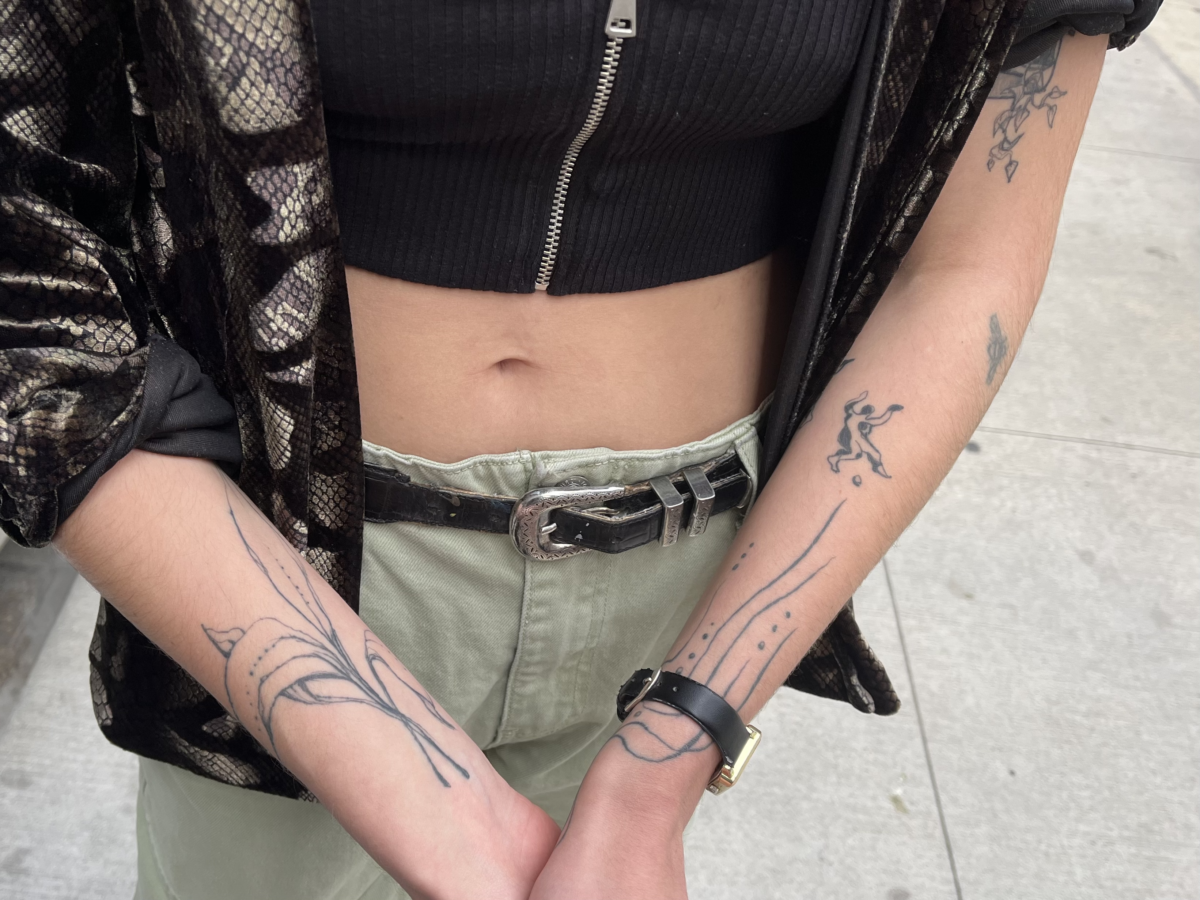 Torso wearing a black crop top and green cargo pants showing forearms covered in tattoos of plants, lines, and human-like figures.