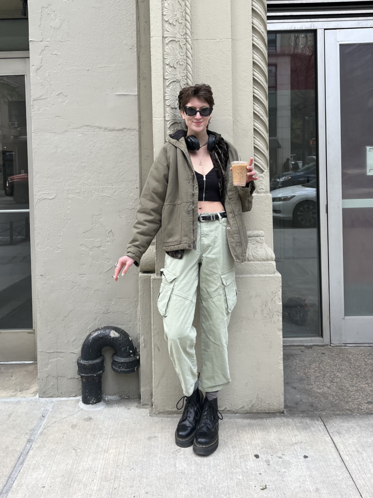 Student wears black boots, green cargo pants, a black crop top, and an army green work jacket while holding an iced coffee and smoking a cigarette in front of a gray building.