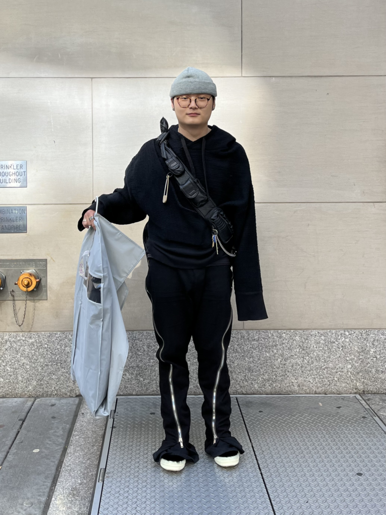 Student wears all-black sneakers, pants with zippers going down the sides, a hoodie, a cross-body bag, and a gray beanie while holding a garment bag in front of a school building on Fifth Avenue.