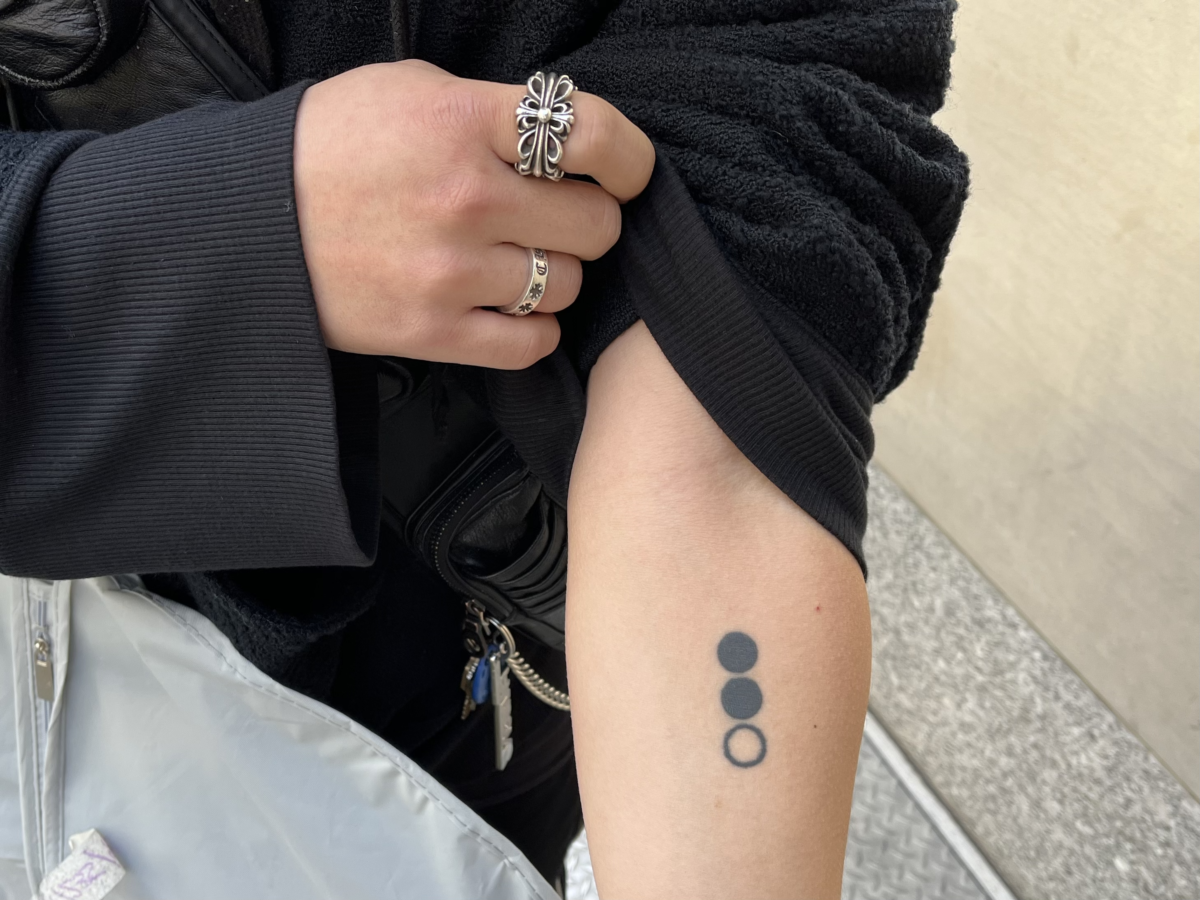 Hand pulls up hoodie sleeve to show a tattoo of two black dots and one white dot in a vertical row.