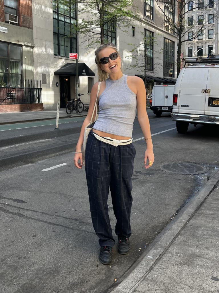 Student wears black sneakers, plaid pants, a gray tank top, black sunglasses, and a tote bag while laughing on Fifth Avenue.