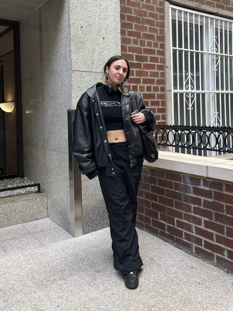 Student wears black sneakers, black cargo pants, black leather jacket, and a black and rhinestone crop top while standing in front of a brick building.