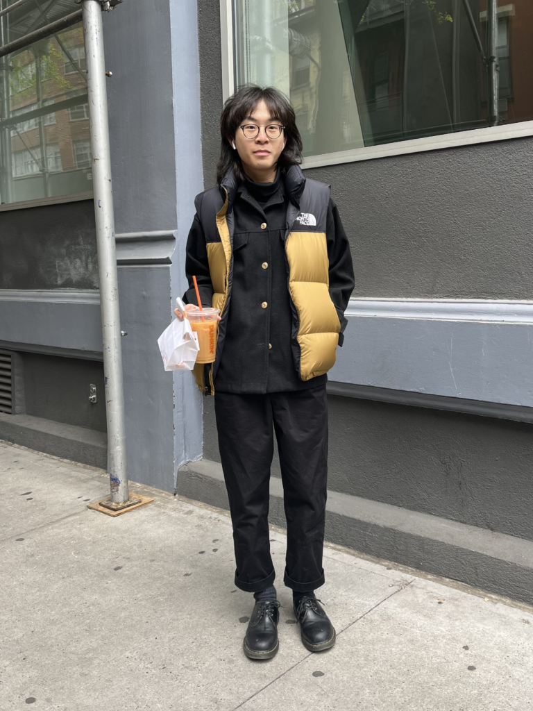 Student wears black boots, black trousers, a black collared shirt, and a camel-colored puffer vest while drinking iced coffee in front of a gray building.