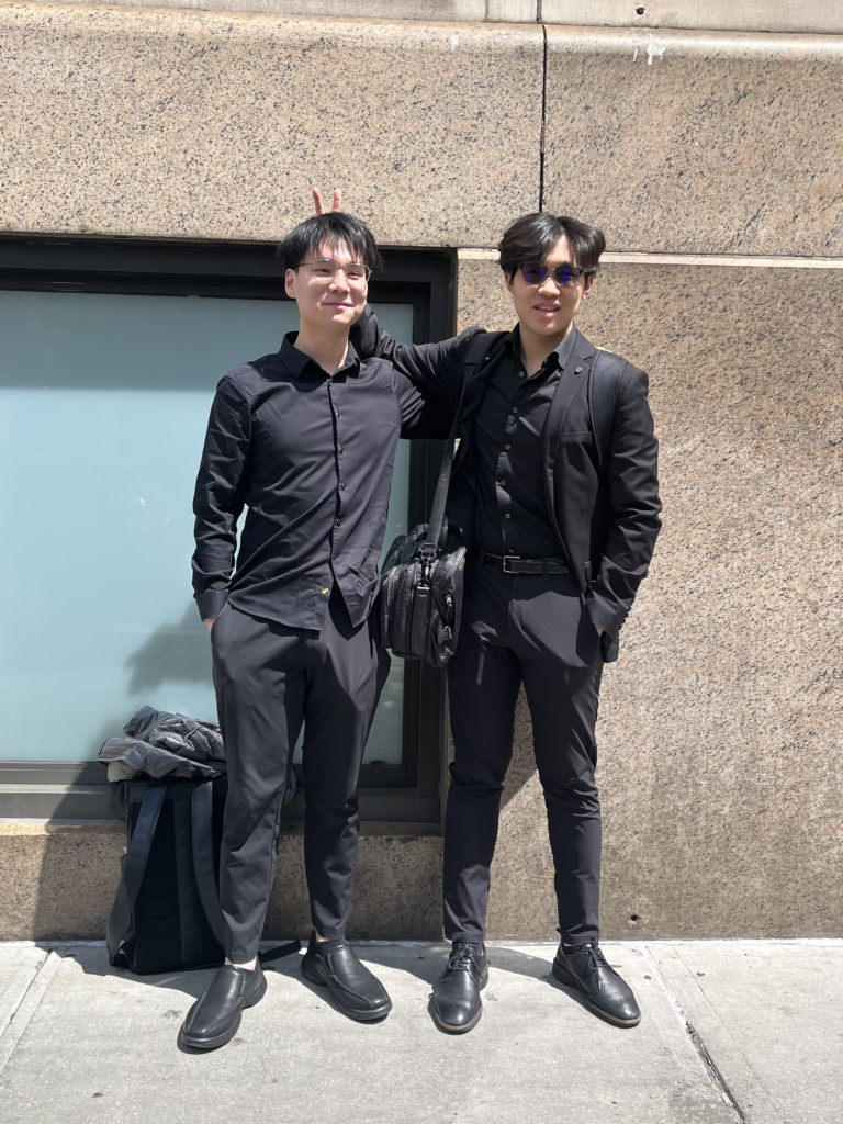 Two students wear all-black suits in front of a beige and blue building. One student holds up a peace sign behind the other’s head.