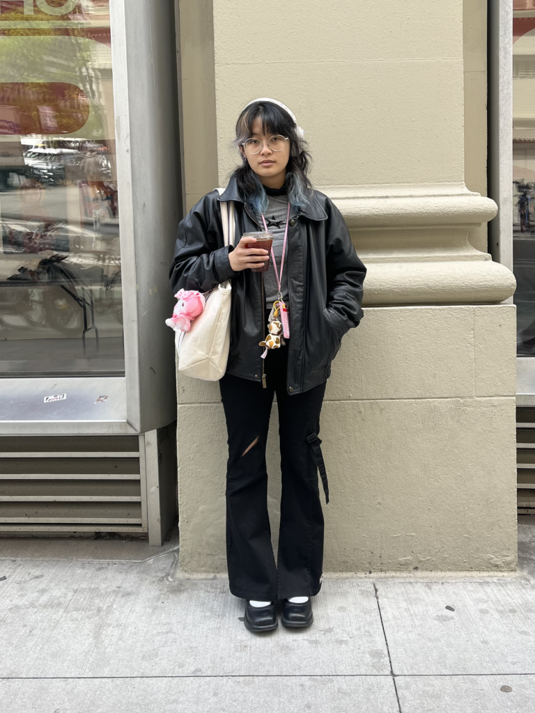 Student wears black loafers, black pants, gray t-shirt, black leather jacket, and a pink lanyard while standing in front of a beige building.