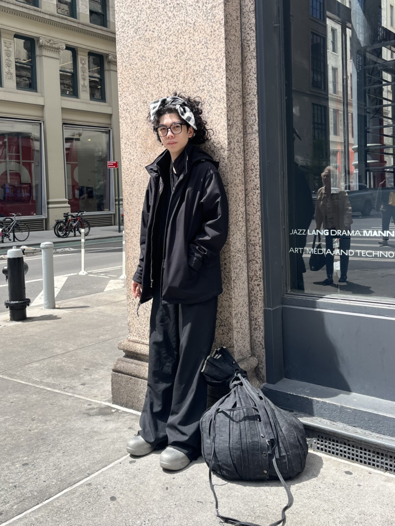 Student wears silver shoes, black pinstripe pants, black jacket, plaid headband, and a gray sack bag while standing in front of a building window.