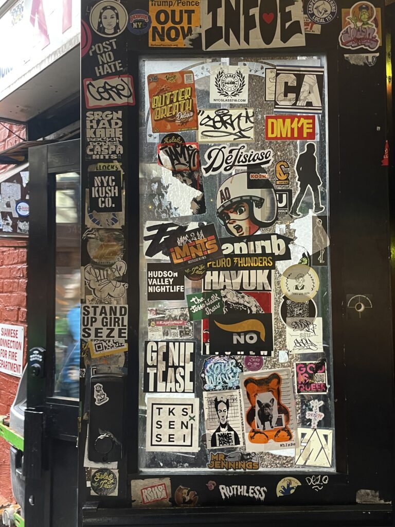 A collage of stickers on an opened black door, as someone exits behind. 