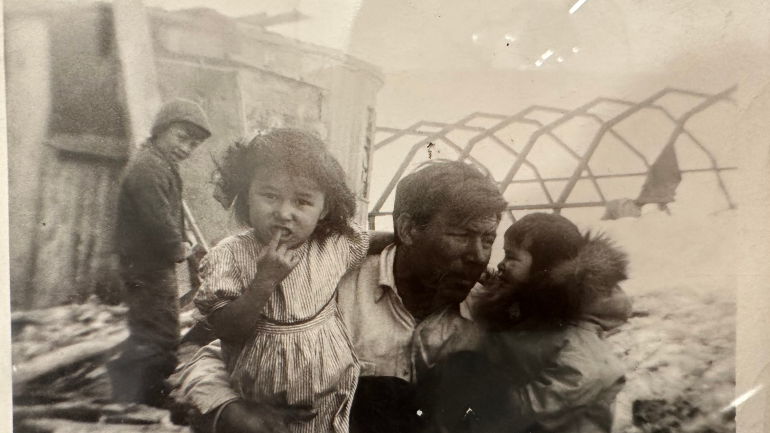 Black and white photo of a Young girl looks into the camera next to her father and sister as they sit in front of their home in Nome, Alaska