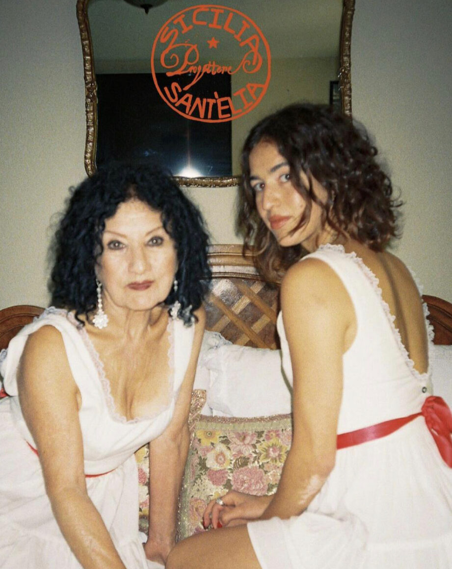 Two women sitting on a bed in front of a mirror in matching white dresses with red ribbons 