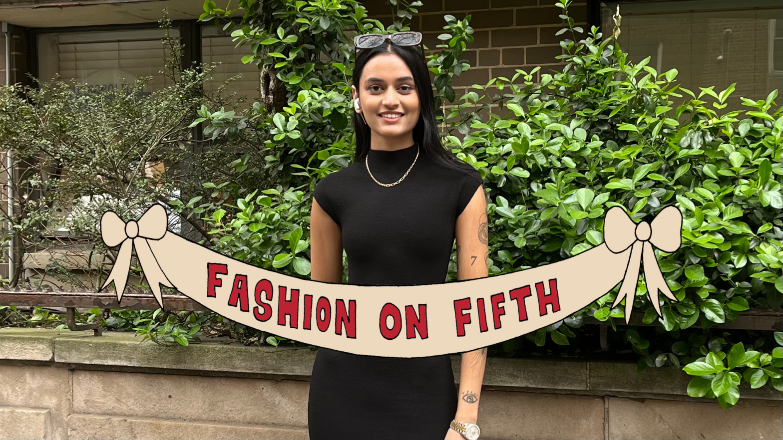 Student wears a short sleeved black dress, gold jewelry, with sunglasses on top of her head, and her left arm is covered in patchwork-style tattoos. She stands in front of green bushes.