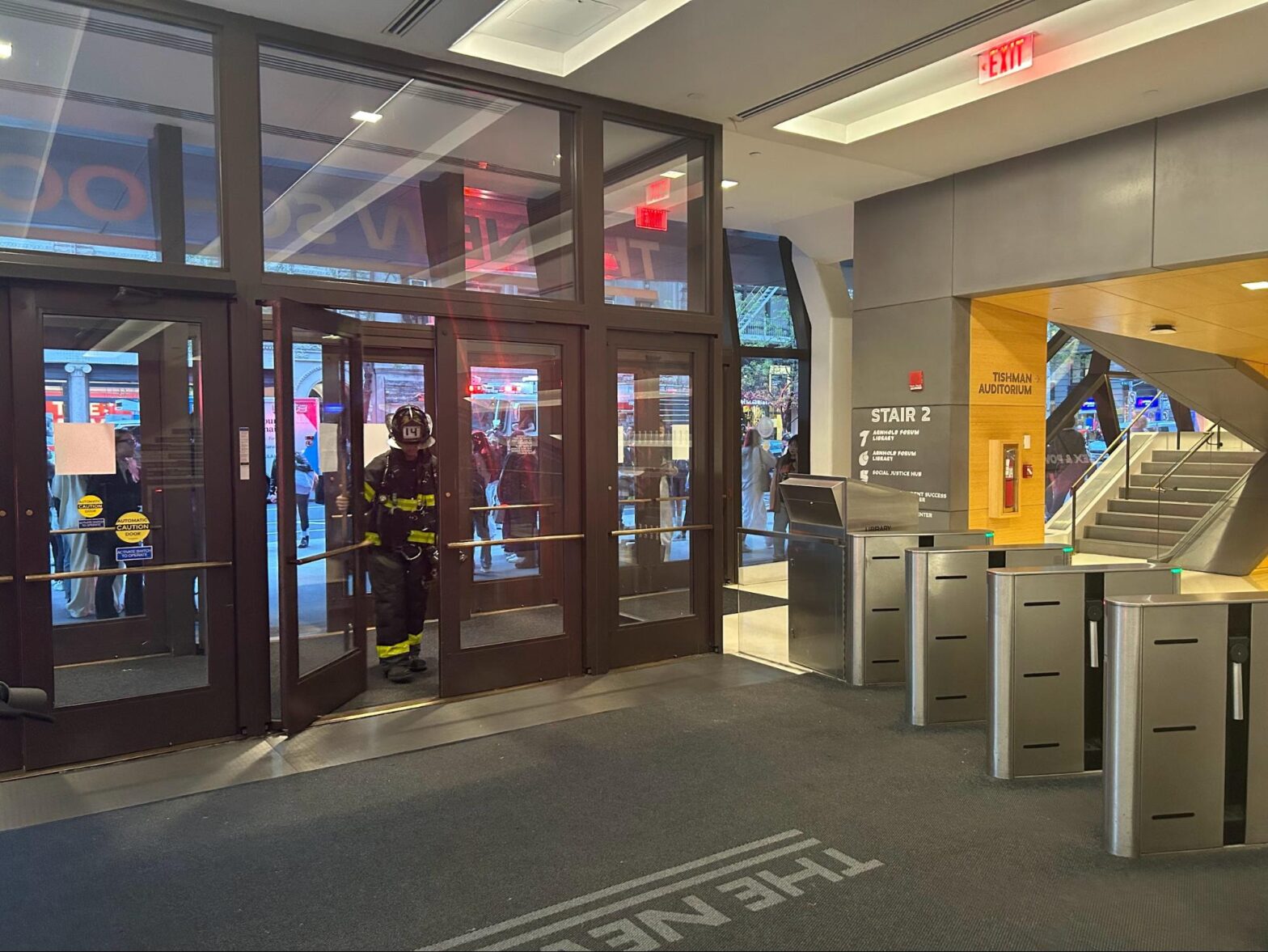 A firefighter dressed in a black and yellow fire suit walks through the glass doors into The New School University Center’s lobby.