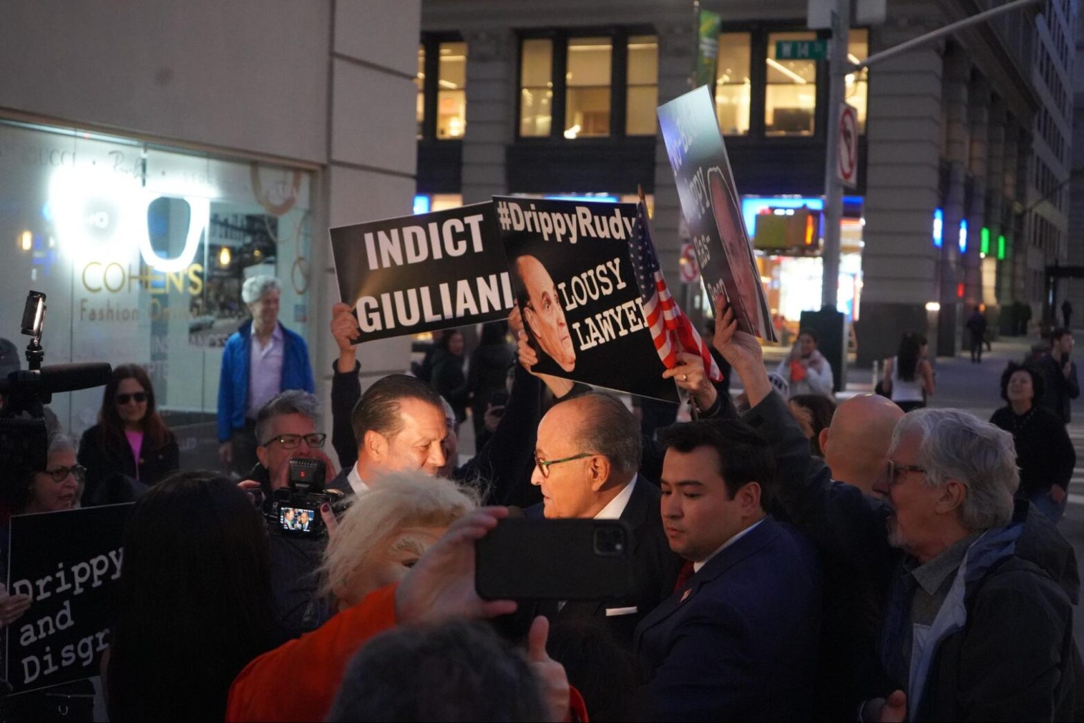 Rudy Giuliani doing an interview while protesters with signs and camera crew surround him.