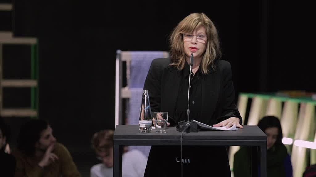 A woman wearing glasses and all black stands on stage with a small desk with a glass of water and a microphone in front of her