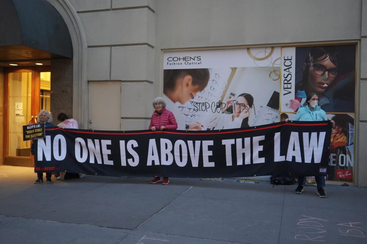 Image of four protestors holding a banner that reads “No One is Above The Law”