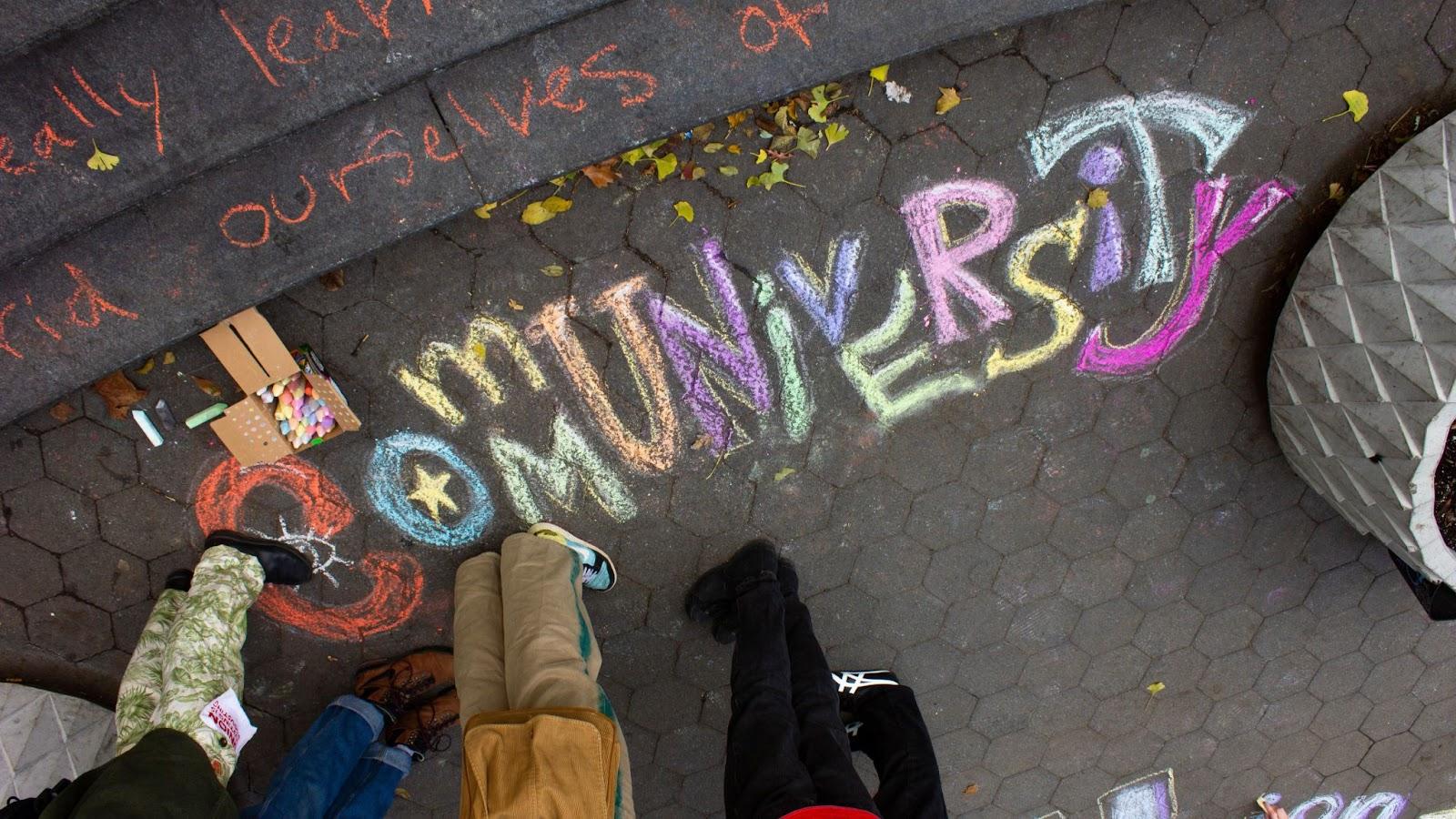 “Communiversity” written in bold letters in colorful chalk on cobblestone. A box of chalk is next to the writing, and a group of people are standing. Only their legs are visible.