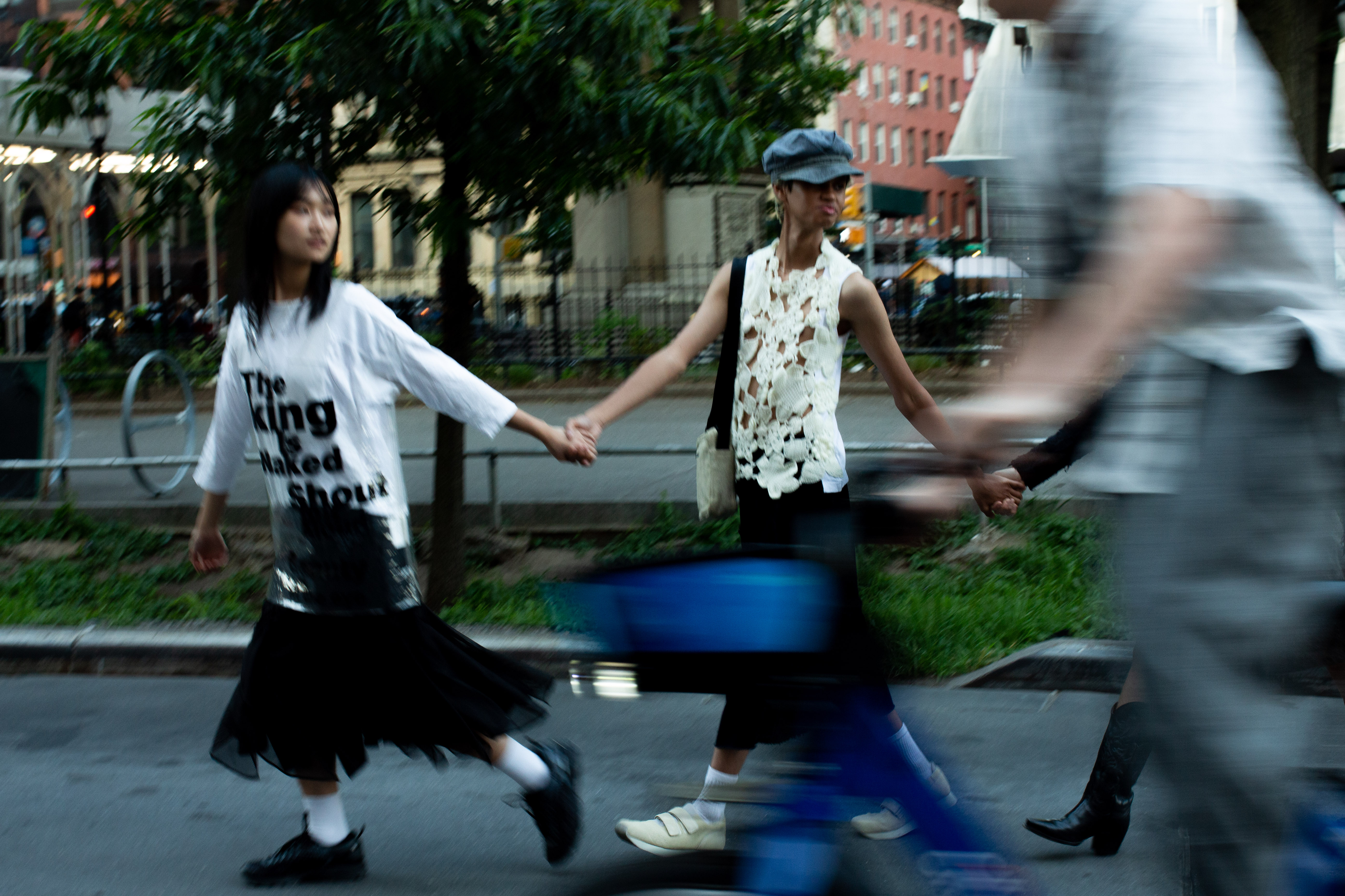 Two people walk hand in hand on pavement in front of a tree with someone riding a bicycle blurred in front of them