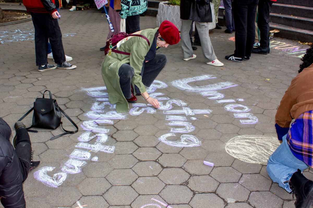 Students kneel down, writing “union busting is disgusting” in purple and white chalk on the cobblestone ground at Washington Square Park