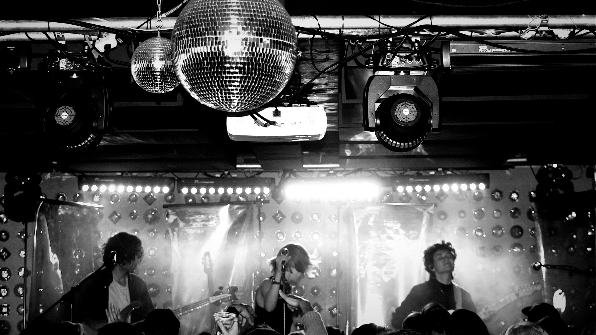 Black and white photo of concert stage. A singer, bassist, and guitarist stand in a line, and a disco ball hangs from the ceiling.