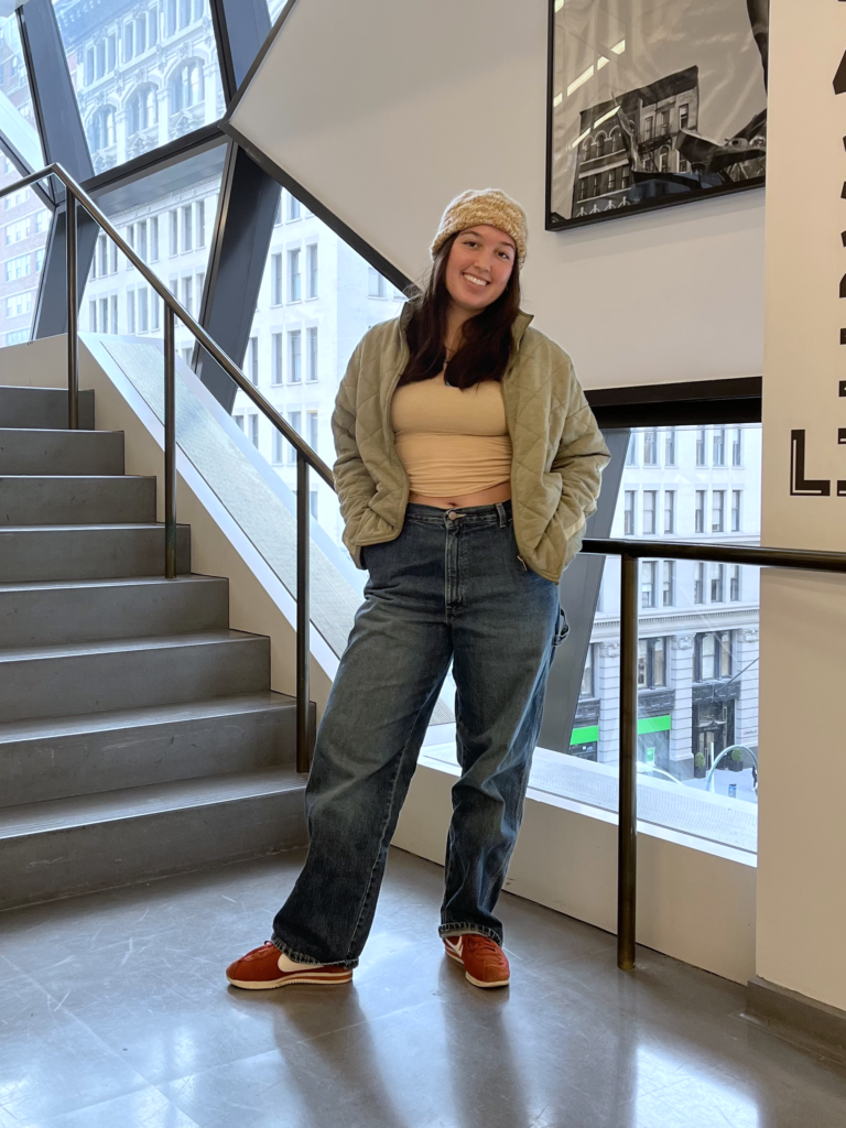 Student wears a beige knit beanie, beige t-shirt, green jacket, blue jeans, and orange Nike Cortez sneakers while standing in front of a staircase.