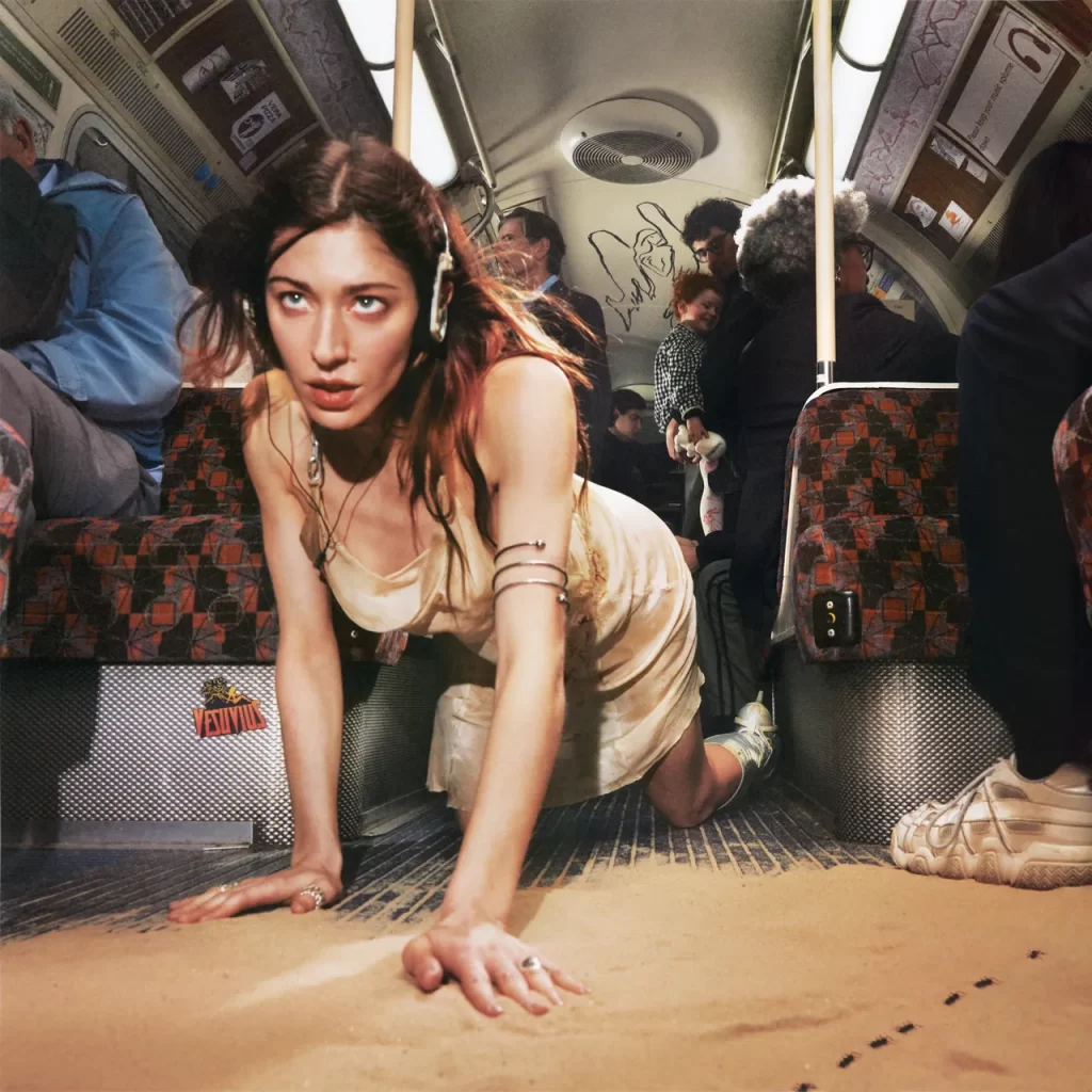Artist Caroline Polachek is on all fours, wearing a retro pair of headphones and a slip dress. She is crawling across the floor of a subway car into a pile of sand. A trail of small insects crawl beside her. Above her are ordinary commuters, and she looks forward with an expression of determination.