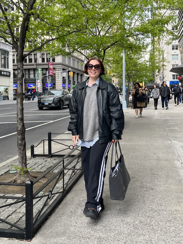 Student wears large sunglasses, a blue button down shirt underneath a gray crewneck sweater, a black leather jacket, striped track pants, and a gray bag while walking down Fifth Avenue.