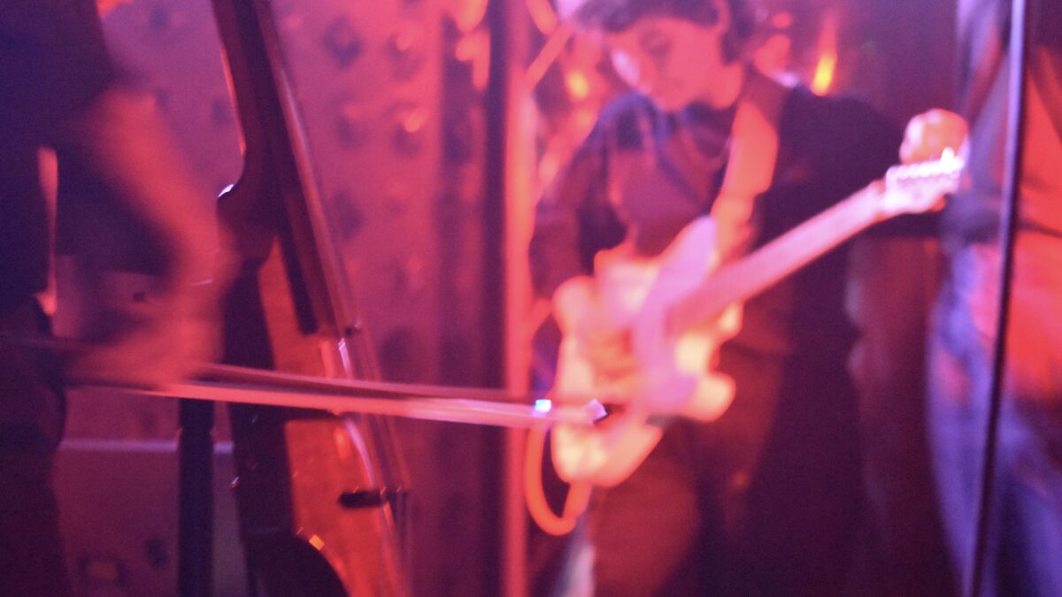 Up close shot features cellist playing cello, with guitarist out of focus in the background. Lighting colors the imagee hues of red and orange. 