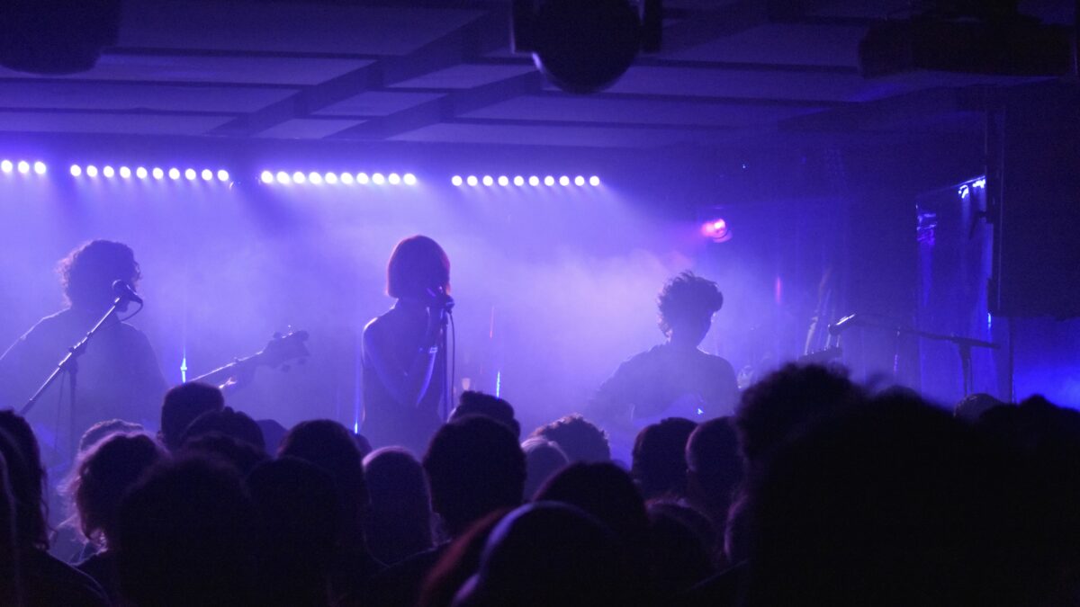 Photo of band perfuming on stage, backlit by purple lights with smoke hazing the image. The tops of the crowds' head is visible. 