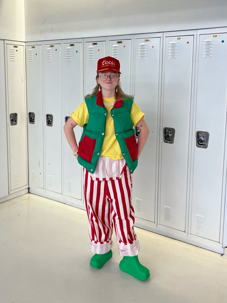 Student wears a red cap, red and green vest, yellow t-shirt, red and white striped overalls, and green Crocs boots with her hands on her hips while standing in front of white lockers.