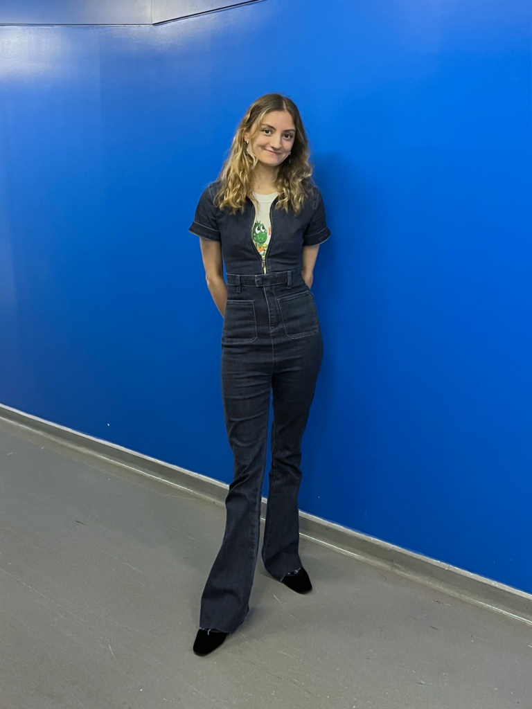 Student wears a white graphic t-shirt, a one-piece denim short sleeved jumpsuit, and black square toe boots while standing in front of a bright blue wall.